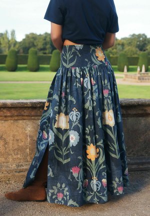 Frau steht im Freien und trägt einen marineblauen Maxirock mit Blumenmuster und seitlichem Schlitz, braune Stiefel und ein schwarzes Oberteil, nahe einer Steinmauer.