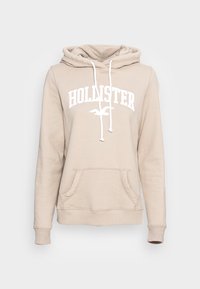 Béžová mikina Hollister s bílými šnůrkami, přední kapsou, ribovanými manžetami a bílým nápisem "HOLLISTER" a logem ptáka na hrudi.