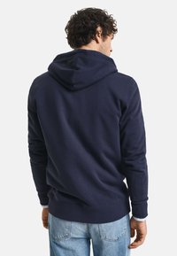 Felpa navy blue con cappuccio regolabile, texture morbida e polsini a costine. Indossata con jeans blu chiaro, mostrando un design semplice e informale.