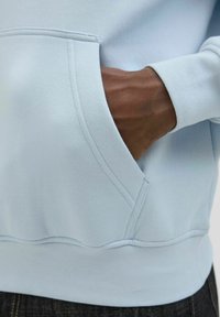 Jasnoniebieska bluza z kangurzym kieszonką, prążkowanymi mankietami i pasem, o miękkiej fakturze materiału oraz widocznymi detalami szwów.