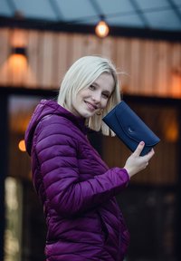 Navy blue wallet with a curved flap, featuring a logo. Woman in a purple puffer jacket holds it, with blurred outdoor lighting in the background.