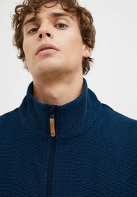 Mørkeblå hettejakke med glidelås i struktureret stof, høy krage og detalj med tre-glidelås. Enkelt design, uformell stil og slim fit.