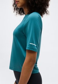 Γυναίκα με σγουρά μαλλιά που φοράει τιρκουάζ μπλουζάκι με κοντά μανίκια και ετικέτα "Προστασία υπεριώδους ακτινοβολίας 50+" στο μανίκι, συνδυασμένο με μαύρο κάτω μέρος.