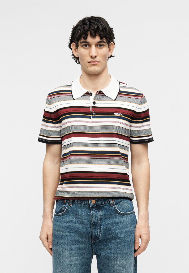 Young man with dark curly hair wearing a short-sleeve, multicolored horizontal striped polo shirt and blue jeans, standing against plain background.