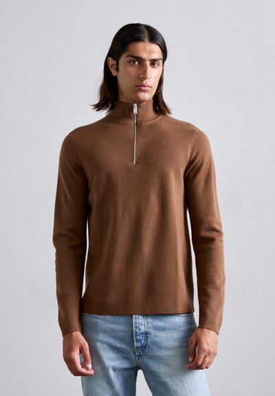 Brauner Strickpullover mit Reißverschluss und langen Ärmeln, weicher Textur und minimalen Designdetails, kombiniert mit hellblauen Denim-Jeans.