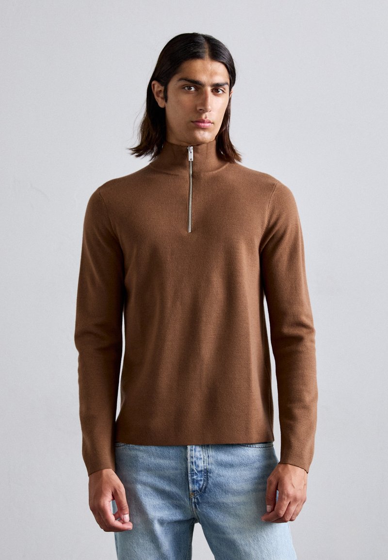 Brauner Strickpullover mit Reißverschluss und langen Ärmeln, weicher Textur und minimalen Designdetails, kombiniert mit hellblauen Denim-Jeans.
