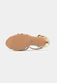 Semelle plate d'une sandale dorée à lanières avec une semelle extérieure en caoutchouc texturé de couleur beige et une bride à la cheville avec boucle sur fond blanc.