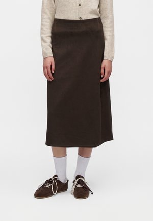 Personne portant un haut manches longues beige avec un bouton, une jupe marron foncé longueur genou, des chaussettes blanches et des baskets marron foncé avec des lacets épais.