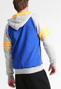 Sudadera con capucha de tela azul con mangas grises y acentos amarillos, incluyendo tres franjas en cada manga. Textura suave.