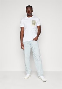 Hvit bomulls-T-skjorte med en grønn firkantet logo. Modellen har på seg lyseblå denimjeans og hvite joggesko, stående mot en grå bakgrunn.