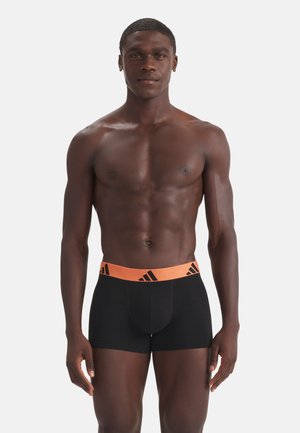 Muskuløs mand stående, vendt fremad iført sorte boxerbriefs med orange taljebånd med sorte Adidas-logoer.