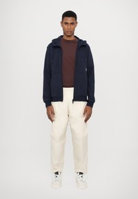 Jūrų laivyno kapučinas su priekinėmis kišenėmis, uždėtas ant rudos marškinėlių, derinamas su grietinėlės spalvos kelnėmis ir baltomis sportbačiais. Lygus audinio paviršius.