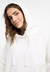 Young woman with wavy hair wearing a white hoodie with a hood, smiling softly against a neutral background.
