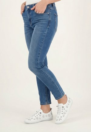 Schmal geschnittene blaue Jeans, kombiniert mit weißen Freizeitschuhen mit Blumenmuster, Hände in den Taschen, stehend vor einem schlichten hellen Hintergrund.