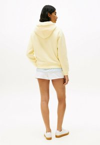 Felpa giallo chiaro con cappuccio posteriore, abbinata a pantaloni corti bianchi. Il tessuto sembra morbido, con una vestibilità comoda e polsini a costine.