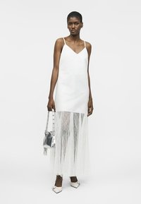 Γυναίκα που φοράει λευκό φόρεμα με λεπτό δαντελένιο φουστάνι, κρατώντας ασημένια τσάντα και φορώντας λευκά μυτερά παπούτσια με φιόγκους.