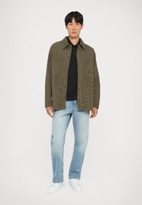 Camicia overshirt in corduroy verde oliva con chiusura a bottoni, due tasche frontali, indossata sopra una polo nera, abbinata a jeans azzurri e sneaker bianche.