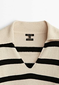 Beige villakashmirneule mustilla vaakasuorilla, jossa on ribbikaarroke ja V-päänmuoto. Lapuissa näkyy tekstiä kauluksessa.