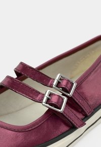 Tumši sarkans satīna apavi ar diviem regulējamiem sudraba sprādzes un noapaļotu purngalu. Zole ir melna ar krēmkrāsas malu, iekšpuse ir gluda.