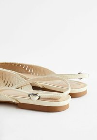 Sandales plates en cuir beige avec des découpes, des sangles à la cheville avec des boucles noires et des semelles marron clair sur un fond blanc.