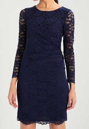 Sømandsblå blondekjole med lange ærmer, der har et blomstermønster og en scalloped kant. Fremstillet af struktureret blonde stof, tætsiddende silhuet.