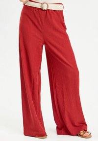 Pantalon rouge à jambes larges en tissu texturé avec une ceinture crème tissée et une boucle circulaire marron. Le design comprend un côtelé vertical.
