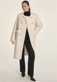 Kremowy płaszcz z mieszanki wełny z detailingiem w postaci złotych guzików, długimi rękawami i luźnym krojem, noszony na czarnym golfie i dopasowanych spodniach.