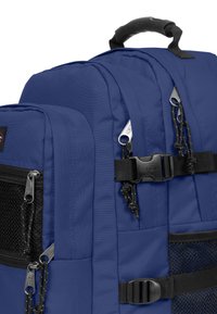 Blå ryggsäck med vadderat tyg, flera dragkedjefickor, nätdetaljer och justerbara axelremmar. Har svarta metalldelar och ett bärhandtag.