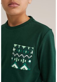 Cămașă verde cu mânecă lungă, având un buzunar cu model în nuanțe de verde și alb, cu desene geometrice, confecționată din material de bumbac moale.