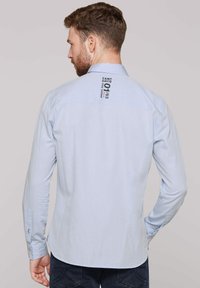 Hellblaues langärmliges Hemd mit strukturierter Oberfläche. Verfügt über einen Umlegekragen und schwarze Druckdetails auf der Rückseite nahe dem Hals.