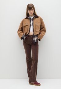 Braune kurze Jacke über einem karierten Hemd und einem weißen Oberteil, kombiniert mit braunen Schlaghosen und kastanienbraunen Turnschuhen, in einem minimalistischen Umfeld.