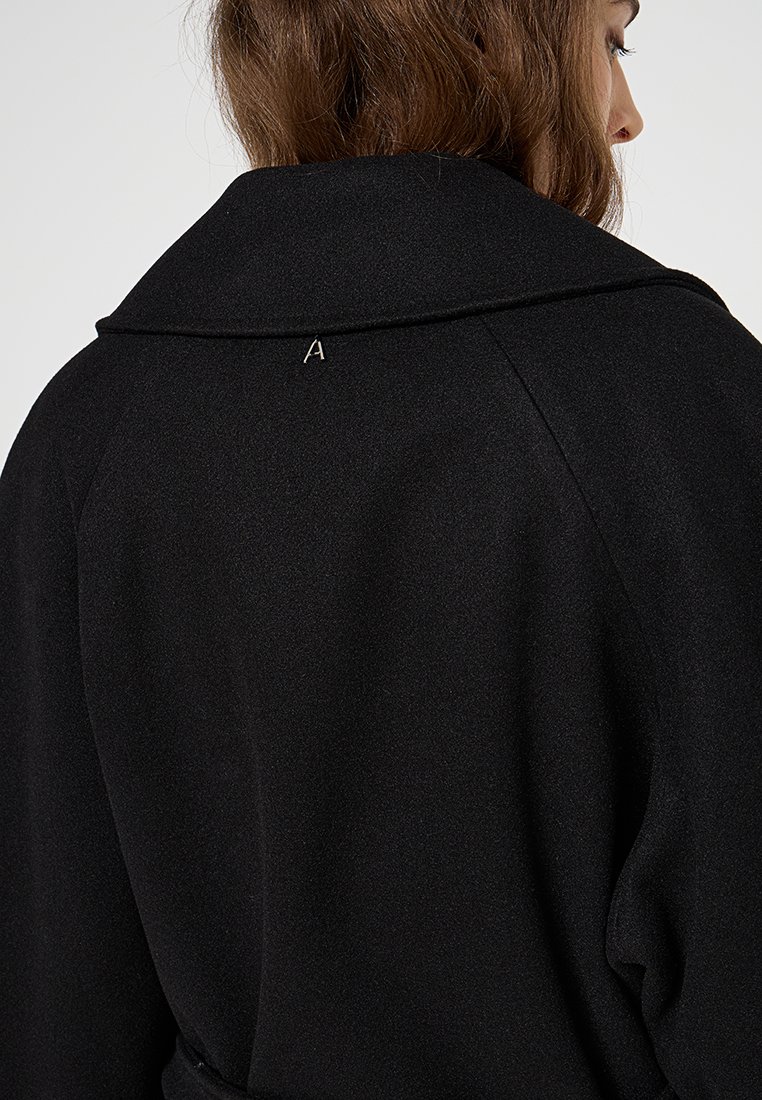 Kobieta z brązowymi włosami, nosząca czarny płaszcz z małym srebrnym napisem "A" na kołnierzu.