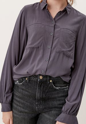 Vrouw draagt een paarse blouse met lange mouwen en knopen, ingestopt in een hoog getailleerde zwarte verweerde spijkerbroek met voorzakken en knoopsluiting.