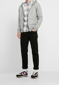 Uomo che indossa una felpa grigia con zip, una camicia a quadri grigia e bianca con bottoni, pantaloni neri arrotolati e sneaker nere, bianche, rosse e blu.