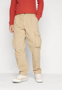 Pantalones cargo color caqui con bolsillos laterales, fabricados con tejido resistente y un corte relajado, combinados con zapatillas blancas y un suéter rojo de canalé.