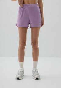 Fioletowe szorty sportowe z elastycznym paskiem, gładką fakturą i bocznymi rozcięciami. W połączeniu z białymi skarpetkami do kostek i srebrnymi trampkami.