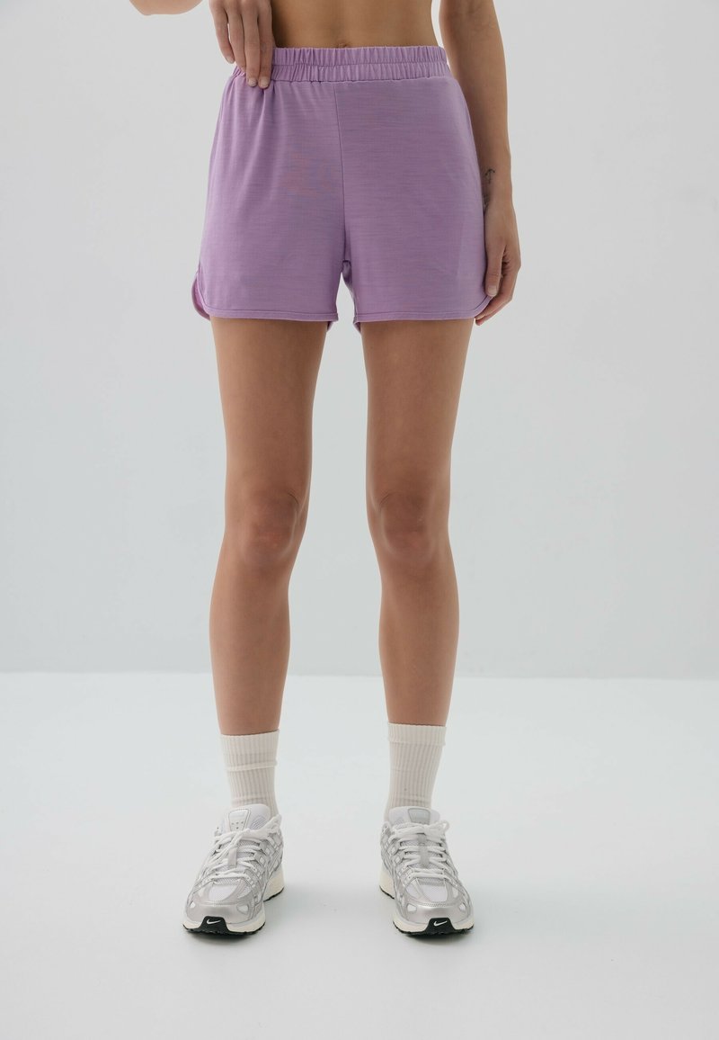 Fioletowe szorty sportowe z elastycznym paskiem, gładką fakturą i bocznymi rozcięciami. W połączeniu z białymi skarpetkami do kostek i srebrnymi trampkami.