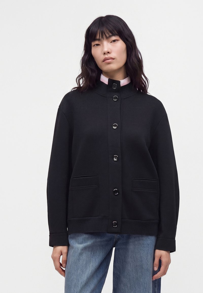 Femme aux cheveux foncés mi-longs porte un cardigan noir boutonné sur une chemise rose clair et un jean bleu sur un fond blanc uni.