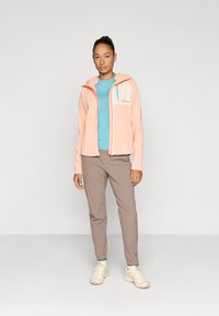 Ανοιχτό ροδακινί μπουφάν με φερμουάρ και μπλε τσέπη, συνδυασμένο με τιρκουάζ πουκάμισο και μπεζ παντελόνι. Το μοντέλο φοράει λευκά αθλητικά παπούτσια.