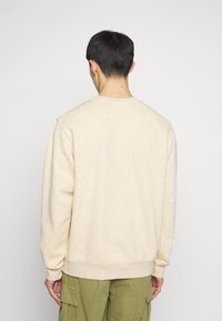 Sweatshirt beige clair en tissu texturé avec des éclats. Il a un col rond et des manches longues, porté avec un pantalon cargo olive.