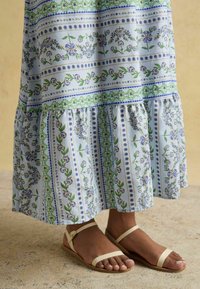 Maxi šaty z svetlomodrej tkaniny s kvetinovými vzormi a vertikálnymi pruhmi. Spodný okraj je volánkový. Skombinované so svetlými sandálmi na remienky.