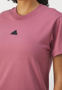 Růžové bavlněné tričko s kulatým výstřihem. Na levé části hrudníku má malé černé logo ve tvaru tří pruhů. Krátké rukávy.