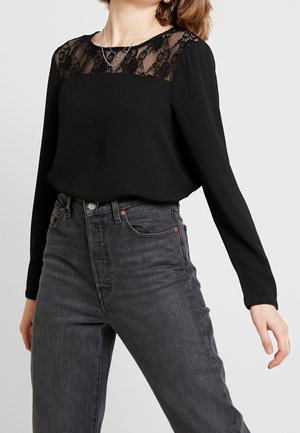 Kobieta w czarnej bluzce z długim rękawem z koronkowym detalem na górze, w połączeniu z ciemnoszarymi jeansami z wysokim stanem i warstwowymi srebrnymi naszyjnikami.