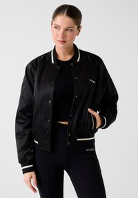 Giacca bomber in satin nero con polsini e collo a coste bianchi a righe, dotata di chiusura a scatto e tasche laterali. Logo del marchio visibile.
