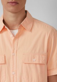 Korte mouwen, licht perzikkleurig knoopshirt met een gestreepte textuur, twee voorzakken en witte knopen.