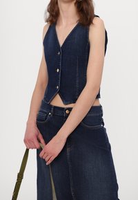 Džínová vesta s výstřihem do V, bez rukávů, s předními knoflíky a zkráceným střihem; doplněná sukní z denimu s vysokým pasem v ladící barvě.