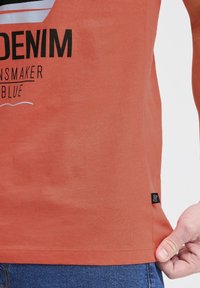 Tricou coral din bumbac moale, cu un imprimeu grafic ce include text și valuri. Eticheta de pe tivul lateral este vizibilă, asortat cu blugi albaștri.