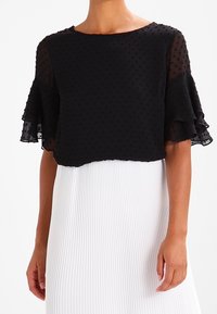 Blusa nera a maniche corte con texture a pois, scollo rotondo e maniche volants, abbinata a una gonna bianca plissettata.