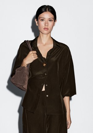 Donkergroene oversized shirt met knoopsluiting, gedragen over een bijpassende outfit. Zachte textuur. Houdt een bruine suède tas met ronde hoeken vast.