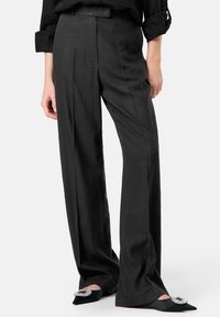 Schwarze Leinenhose mit weitem Bein, hoher Taille und sichtbaren Falten. Kombiniert mit schwarzen spitzen Schuhen, die mit einem Schmuckakzent verziert sind.